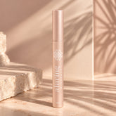 A single cylindrical cosmetic tube standing upright on a beige surface, with warm sunlight and leaf shadows cast across the scene. The tube is metallic blush pink with white printed branding reading 'alluring minerals' and a floral emblem near the top.