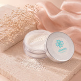 An open round metal tin of Alluring Minerals powder sits beside its lid on a beige stone slab, with a clear scoop inside and a fine white powder spilled out. The scene is styled with soft peach fabric, dried baby's breath flowers, and a textured stone background.
