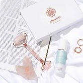 Alluring Minerals skincare set featuring a rose gold and pink stone facial roller, a pink stone gua sha, a white and mint green serum bottle, and two rose gold rings, styled on white fabric and an open book next to a branded white gift box.