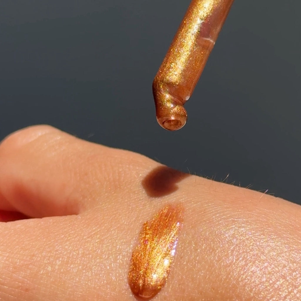 A close-up of a metallic gold shimmer cosmetic product on a person's skin, with a dropper above showing the reflective golden liquid.