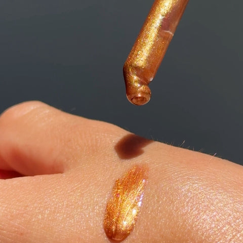 A close-up of a metallic gold shimmer cosmetic product on a person's skin, with a dropper above showing the reflective golden liquid.