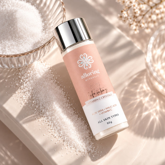 A bottle of alluring minerals Vitamin C Crystals skincare product, styled on a beige tray with scattered crystals, a bowl of white sugar, and dried flowers under warm, direct sunlight.